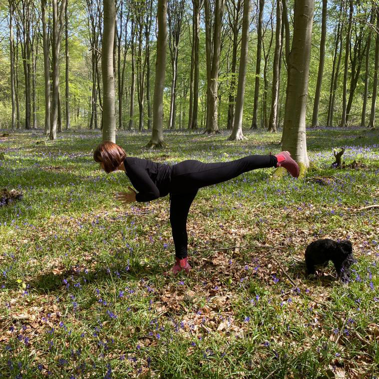 Jane balancing in warrior three among bluebells in a beech wood